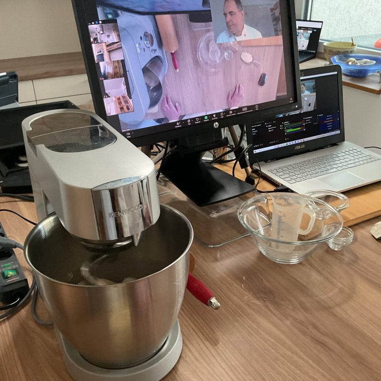 Küchenmaschine steht auf einem Tisch neben einem Laptop mit Kochvideo und Zutaten.